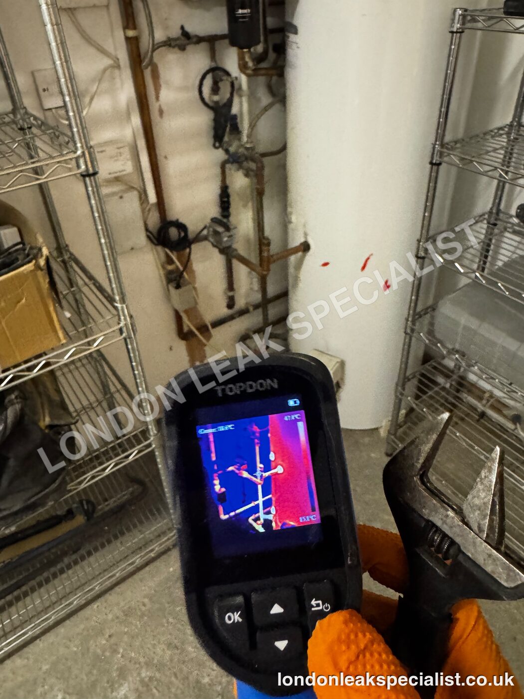 A thermal imaging camera screen held in front of boiler pipework and a hot water cylinder in a utility space