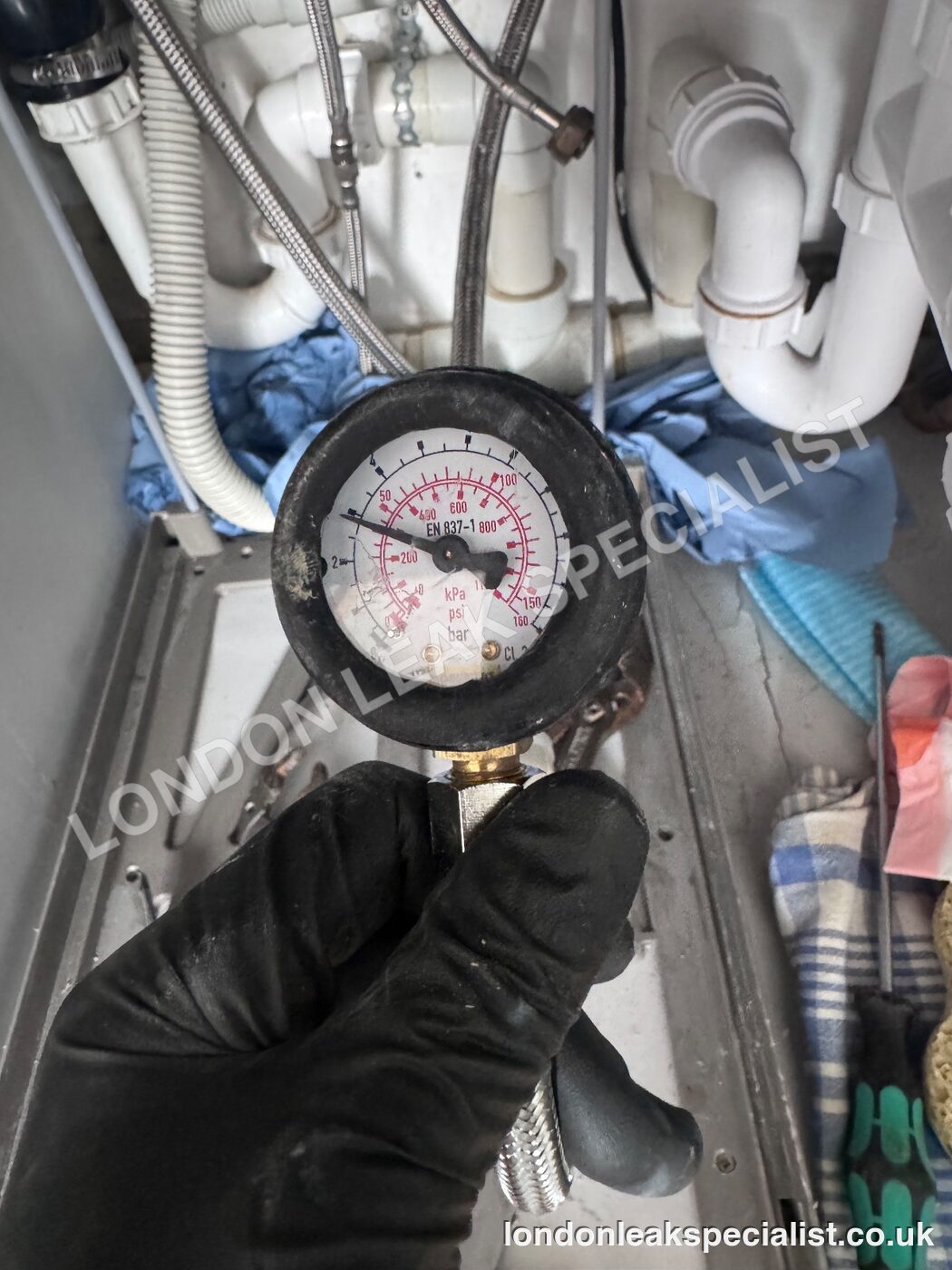 A pressure gauge held in a black glove during a water pressure test, with pipework visible behind
