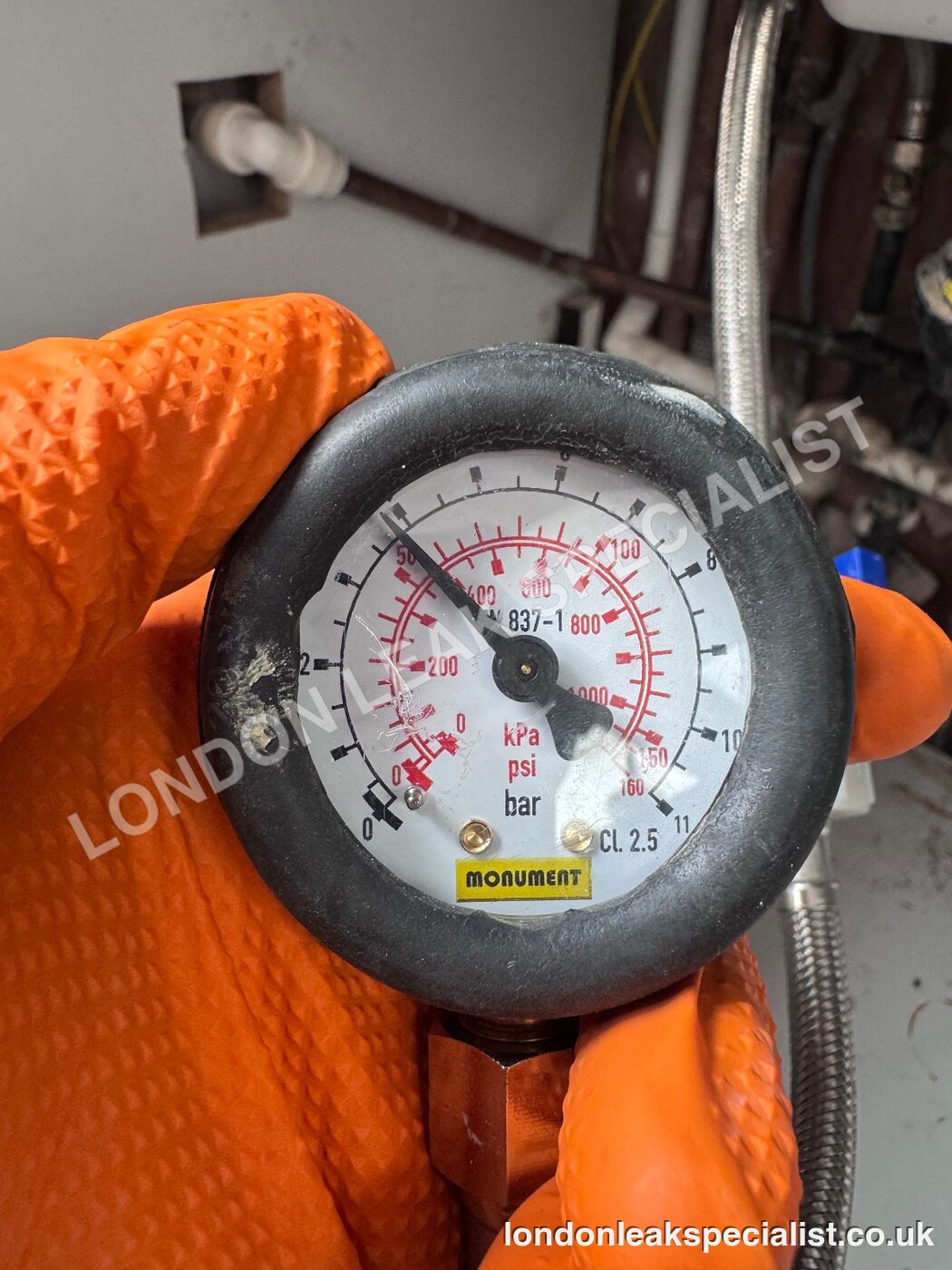 A pressure gauge held in an orange glove during a cold water pressure test beneath a kitchen sink