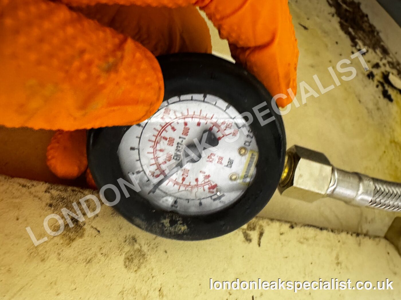 A hand in an orange glove holding a pressure gauge connected to a flexible hose during a supply pressure test